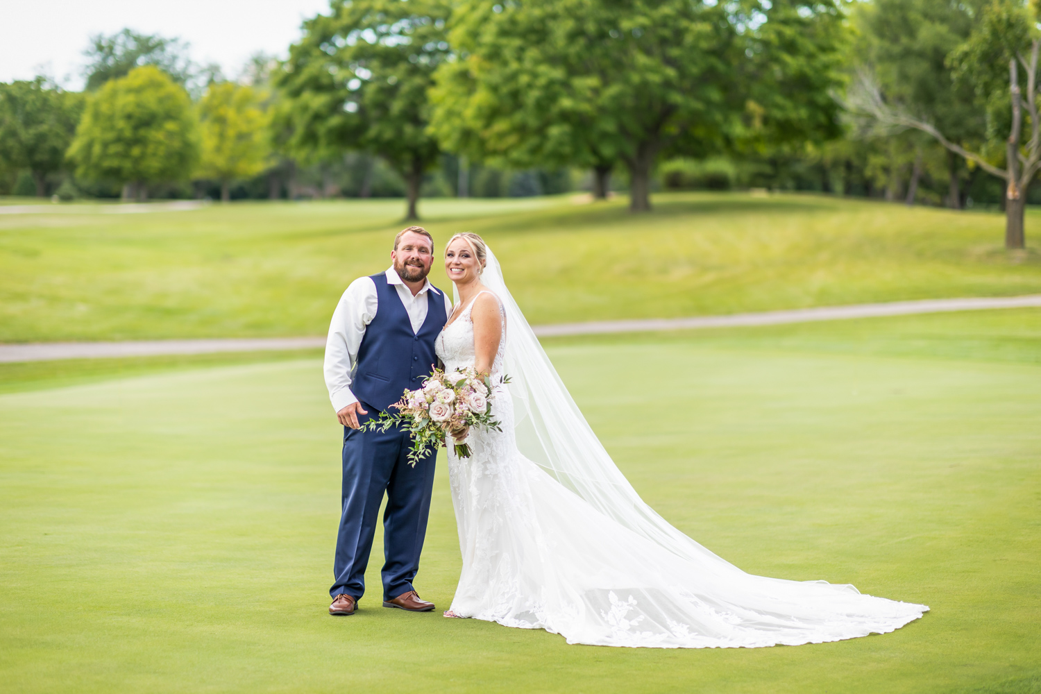 A new couple posing on their wedding day on a field of greens with trees in the background.